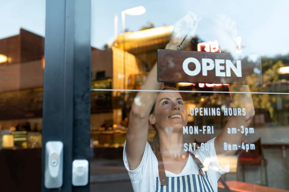 A smiling woman hanging an OPEN sign in her shop window.